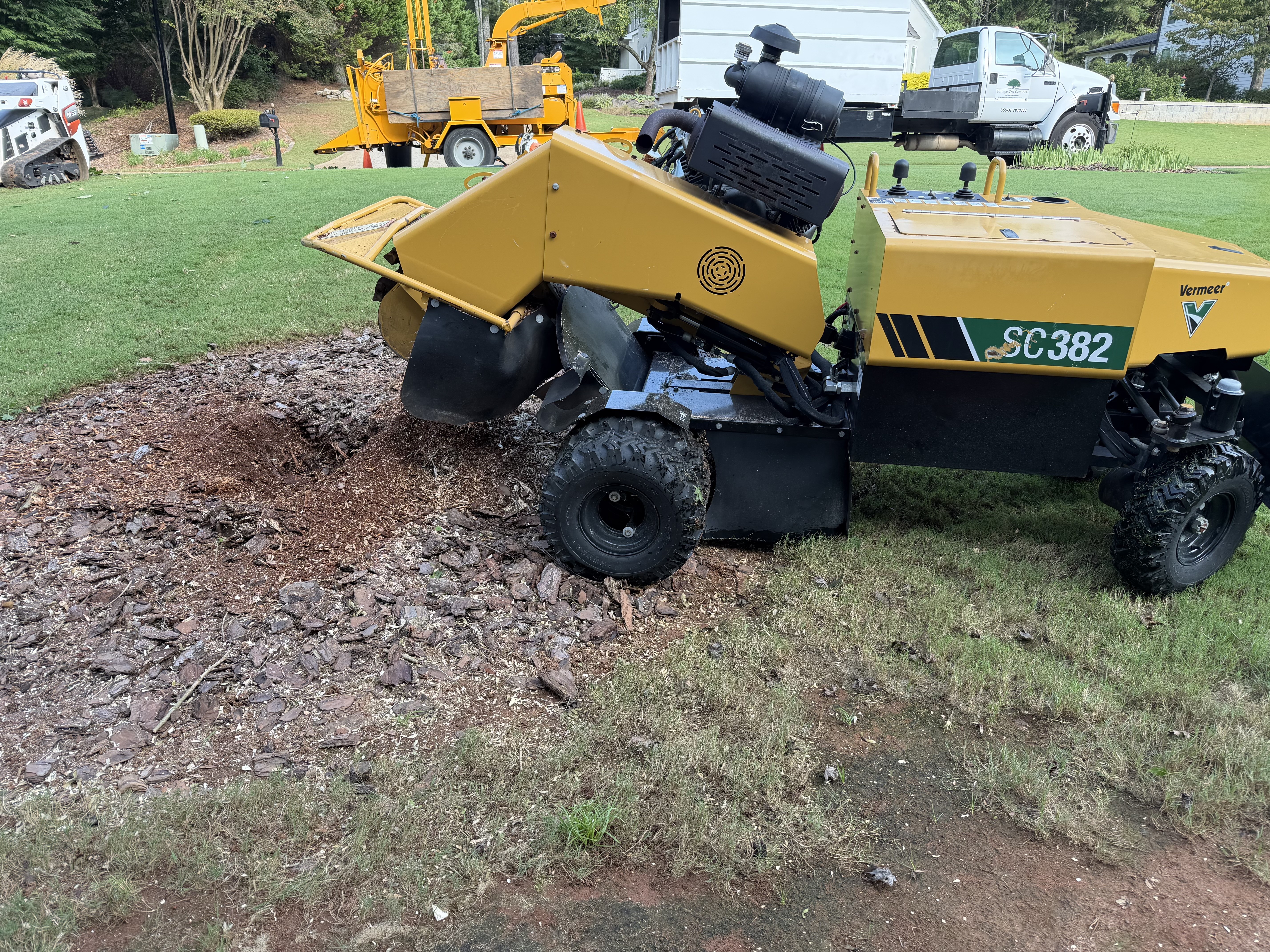 Heritage Tree Care’s yellow Vermeer SC382 stump grinder beside a ground stump, with the crew’s truck and equipment behind it